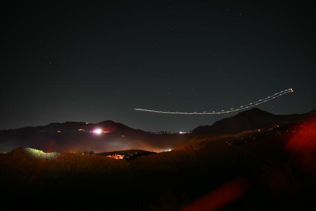 This long-exposure photograph shows the light trail of a helicoper dropping water over the Bryson Fire in Malibu, California on October 15, 2026. The brush fire in the mountains west of Los Angeles prompted evacuation warnings from officials, as powerful Santa Ana winds risked spreading the blaze rapidly. (Photo by Patrick T. Fallon / AFP)