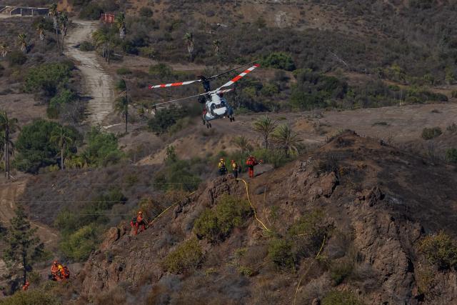 Inmate firefighters walk in a burned hillside looking for hotspots while a helicopter flies over the Bryson Fire in Malibu, California on October 16, 2026. A brush fire in the mountains west of Los Angeles prompted evacuation warnings from officials on Thursday, as powerful winds risked spreading the blaze rapidly. (Photo by Apu GOMES / AFP)
