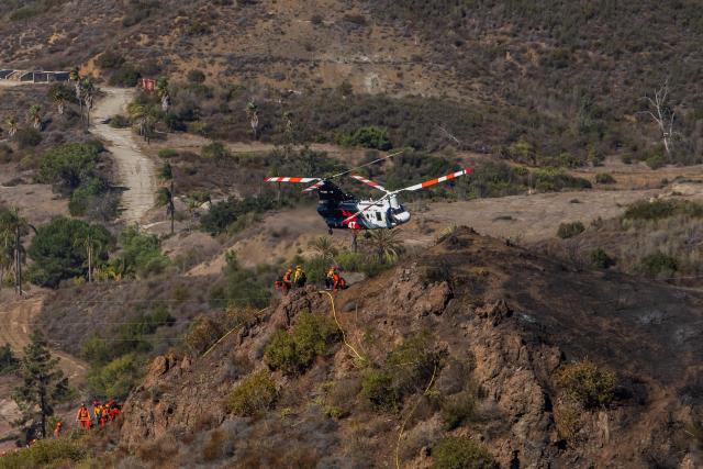 Inmate firefighters walk in a burned hillside looking for hotspots while a helicopter flies over the Bryson Fire in Malibu, California on October 16, 2026. A brush fire in the mountains west of Los Angeles prompted evacuation warnings from officials on Thursday, as powerful winds risked spreading the blaze rapidly. (Photo by Apu GOMES / AFP)