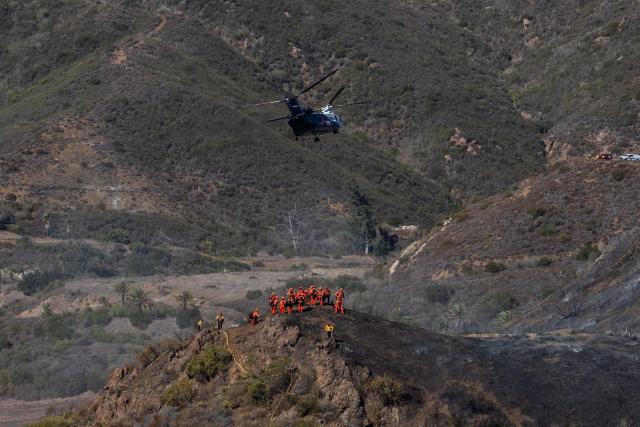 Inmate firefighters walk in a burned hillside looking for hotspots while a helicopter flies over the Bryson Fire in Malibu, California on October 16, 2026. A brush fire in the mountains west of Los Angeles prompted evacuation warnings from officials on Thursday, as powerful winds risked spreading the blaze rapidly. (Photo by Apu GOMES / AFP)