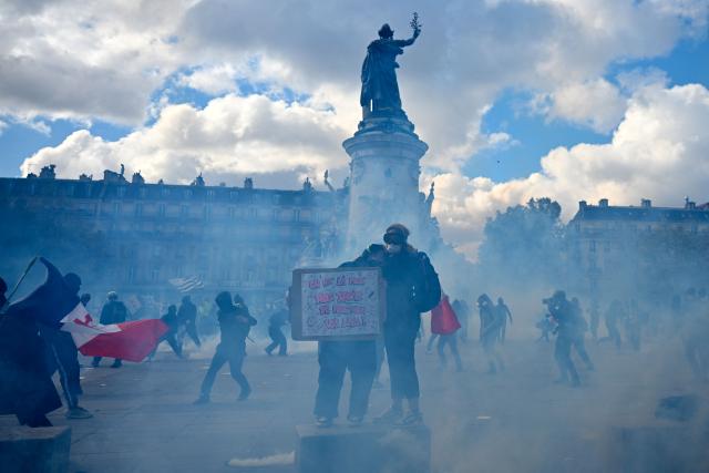 A protester holds a placard amid tear gas smoke as clashes erupt ahead of a demonstration to protest against the "high cost of living", at Place de la Republique in Paris on October 17, 2026 as part of a nationwide call for mobilisation bringing together various groups expressing their anger: former "Yellow Vests", secondary school and university students, climate activists and even farmers. After weeks of student protests, France faces broader, nationwide demonstrations on October 17, 2026 over the cost of living, setting up a potentially fraught six months heading into a presidential election. A police source told AFP that authorities estimated nearly 200,000 people could take part, with 40,000 in Paris. (Photo by Matthieu RONDEL / AFP)