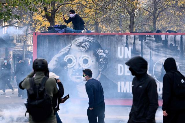 Protesters climb on an billboard amid tear gas smoke as clashes erupt ahead of a demonstration to protest against the "high cost of living", at Place de la Republique in Paris on October 17, 2026 as part of a nationwide call for mobilisation bringing together various groups expressing their anger: former "Yellow Vests", secondary school and university students, climate activists and even farmers. After weeks of student protests, France faces broader, nationwide demonstrations on October 17, 2026 over the cost of living, setting up a potentially fraught six months heading into a presidential election. A police source told AFP that authorities estimated nearly 200,000 people could take part, with 40,000 in Paris. (Photo by Guillaume Saligot / AFP)