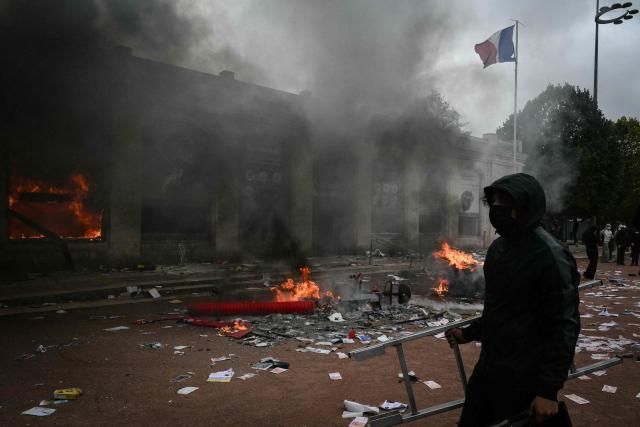 A protester walks past Lyon's tourism office set on fire during a demonstration to protest against the "high cost of living", in Lyon, southeastern France, on October 17, 2026 as part of a nationwide call for mobilisation bringing together various groups expressing their anger: former "Yellow Vests", secondary school and university students, climate activists and even farmers. After weeks of student protests, France faces broader, nationwide demonstrations on October 17, 2026 over the cost of living, setting up a potentially fraught six months heading into a presidential election. A police source told AFP that authorities estimated nearly 200,000 people could take part, with 40,000 in Paris. (Photo by Jeff PACHOUD / AFP)