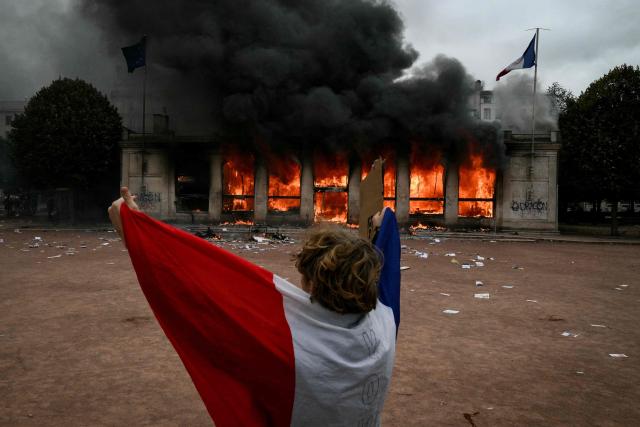 TOPSHOT - A protester wrapped in a French national flag stands in front of Lyon's tourism office set on fire during a demonstration to protest against the "high cost of living", in Lyon, southeastern France, on October 17, 2026 as part of a nationwide call for mobilisation bringing together various groups expressing their anger: former "Yellow Vests", secondary school and university students, climate activists and even farmers. After weeks of student protests, France faces broader, nationwide demonstrations on October 17, 2026 over the cost of living, setting up a potentially fraught six months heading into a presidential election. A police source told AFP that authorities estimated nearly 200,000 people could take part, with 40,000 in Paris. (Photo by Jeff PACHOUD / AFP)