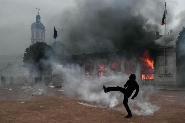 TOPSHOT - A protester kicks a projectile as he stands among tear gas in front of Lyon's tourism office during a demonstration to protest against the "high cost of living", in Lyon, southeastern France, on October 17, 2026 as part of a nationwide call for mobilisation bringing together various groups expressing their anger: former "Yellow Vests", secondary school and university students, climate activists and even farmers. After weeks of student protests, France faces broader, nationwide demonstrations on October 17, 2026 over the cost of living, setting up a potentially fraught six months heading into a presidential election. A police source told AFP that authorities estimated nearly 200,000 people could take part, with 40,000 in Paris. (Photo by Jeff PACHOUD / AFP)