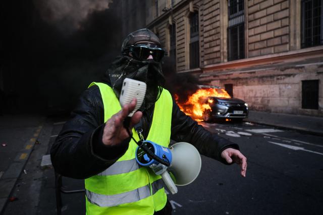 A protester wearing a yellow vest walks past a car on fire during a demonstration to protest against the "high cost of living", in Paris on October 17, 2026 as part of a nationwide call for mobilisation bringing together various groups expressing their anger: former "Yellow Vests", secondary school and university students, climate activists and even farmers. After weeks of student protests, France faces broader, nationwide demonstrations on October 17, 2026 over the cost of living, setting up a potentially fraught six months heading into a presidential election. A police source told AFP that authorities estimated nearly 200,000 people could take part, with 40,000 in Paris. (Photo by Kiran RIDLEY / AFP)