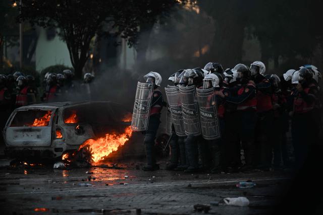 Riot police take cover as they stand in front of a burning car during an anti-government demonstration in central Tirana on October 17, 2026, as protests continued for a fifth consecutive day over university conditions and demands for the resignation of Albanian Prime Minister. Several hundred people, some masked, gathered around noon in front of the ministry of education, with protests later spreading across the city. Protesters threw rocks at the police and set litter bins on fire, using them as barricades to prevent the police from advancing. Armend NIMANI / AFP (Photo by ARMEND NIMANI / AFP)