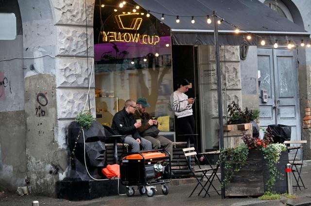 Visitors rest outside a coffee house in Kyiv, near a working power generator during an outage on October 18, 2026, caused by attacks on energy facilities in the country, amid the Russian invasion of Ukraine. (Photo by Sergei SUPINSKY / AFP)