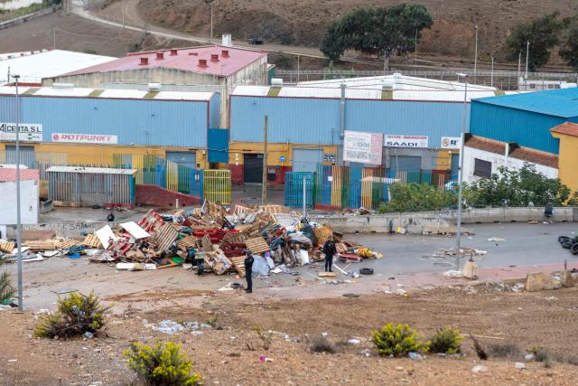 Police forces stand as workers clear debris from a dismantled migrant settlement at the entrance to the Tarajal warehouses, in Spain's North African enclave of Ceuta on October 18, 2026. Scores of migrants remain sleeping rough in outlying neighborhoods of the city since the massive influx of tens of thousands of migrants from Morocco in late July, fueling tension in this Spanish North African enclave of some 80,000 inhabitants. Migrants are being transported by bus to police facilities, with minors taken to the National Police headquarters and adults transferred to the Tarajal Temporary Assistance Centre for Foreigners (CATE) for identification, registration and screening. (Photo by Antonio Sempere / AFP)