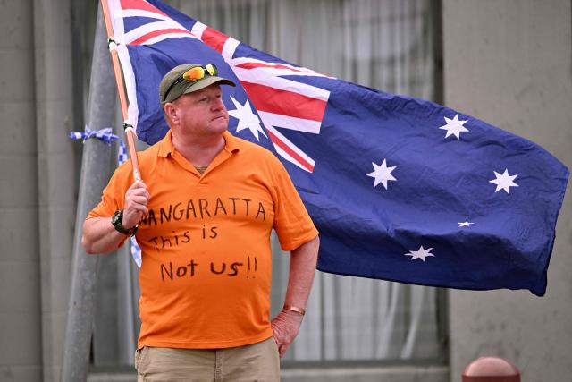 A man holds an Australian flag outside the office of Australian politician Helen Haines in the northern regional city of Wangaratta, about 250 kilometres north-east of Melbourne on October 20, 2026. Australian independent lawmaker Helen Haines underwent surgery overnight but remains in a critical condition after being struck deliberately by a car in an attack, Prime Minister Anthony Albanese said on October 20. Political violence is rare in Australia, but law enforcement have documented a steady rise in threats and abuse directed towards the nation's elected officials in recent years. (Photo by William WEST / AFP)