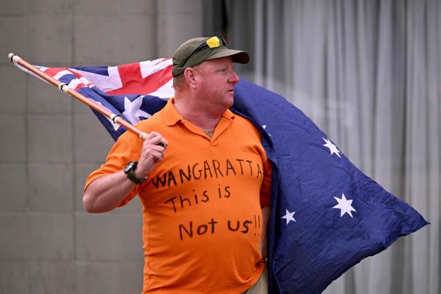 A man holds an Australian flag outside the office of Australian politician Helen Haines in the northern regional city of Wangaratta, about 250 kilometres north-east of Melbourne on October 20, 2026. Australian independent lawmaker Helen Haines underwent surgery overnight but remains in a critical condition after being struck deliberately by a car in an attack, Prime Minister Anthony Albanese said on October 20. Political violence is rare in Australia, but law enforcement have documented a steady rise in threats and abuse directed towards the nation's elected officials in recent years. (Photo by William WEST / AFP)
