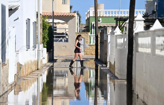 A woman with rubber boots crosses a street following torrential downpours by the beach in Oliva, in the Valencia region, eastern Spain on October 20, 2026. Torrential rain battered eastern Spain, unleashing severe flooding that left at least four people dead in recent days, inundated streets and forced many residents to stay indoors. (Photo by Jose JORDAN / AFP)