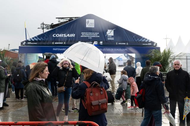 People wait under the rain in front of the entrance of the village of the Route du Rhum, which is closed due to weather conditions, in Saint-Malo, western France on October 21, 2026, a few days prior to the start of the 13th Route du Rhum - Destination Guadeloupe. From Saint-Malo in Brittany to Pointe-a-Pitre in Guadeloupe, over 100 skippers across 6 classes will take the start on November 1, 2026, in this solo transatlantic sailing race. (Photo by Sebastien Salom-Gomis / AFP)