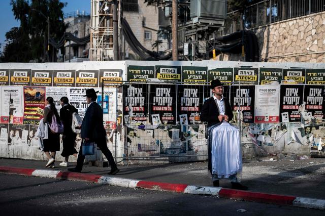 An ultra-Orthodox Jewish man holding his laundry waits in front of torn electoral posters among other posters on a wall along a street in Jerusalem on October 21, 2026, ahead of Israel's legislative election. Israel heads to the polls on October 27 in its first election since the October 7, 2023 Hamas-led attack, in what is seen as a referendum on the prime minister. (Photo by Jewel SAMAD / AFP)