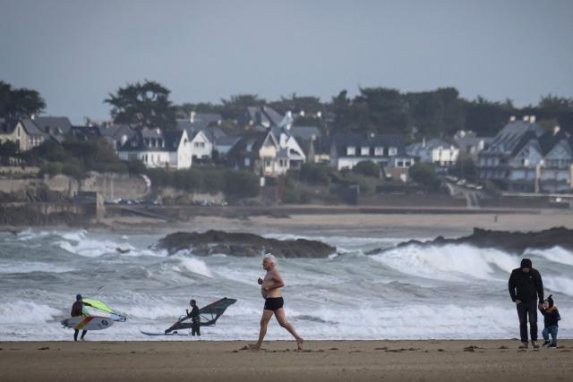 A jogger runs on the sand next to windsurfers (L) and a person standing with a child (R) with waves of the Channel hitting rocks and residential buildings in the background, as the village of the Route du Rhum had been closed due to weather conditions, in Saint-Malo, western France on October 21, 2026, a few days prior to the start of the 13th Route du Rhum - Destination Guadeloupe. (Photo by Loic VENANCE / AFP)