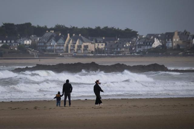 A group of pedestrians walk on the sand of a windy beach with waves of the Channel hitting rocks, as the village of the Route du Rhum had been closed due to weather conditions, in Saint-Malo, western France on October 21, 2026, a few days prior to the start of the 13th Route du Rhum - Destination Guadeloupe. (Photo by Loic VENANCE / AFP)