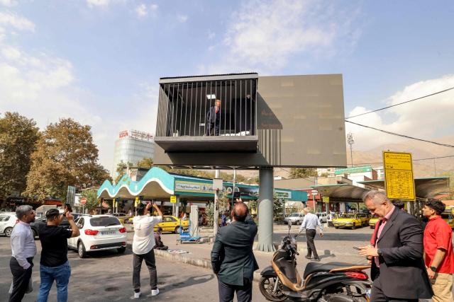 Pedestrians take pictures of an Iranian actor wearing a mask depicting US President Donald Trump as he poses inside a mock prison cell installed onto a street billboard at Tajrish Square in Tehran on October 21, 2026. (Photo by ATTA KENARE / AFP) / ALTERNATIVE CROP
