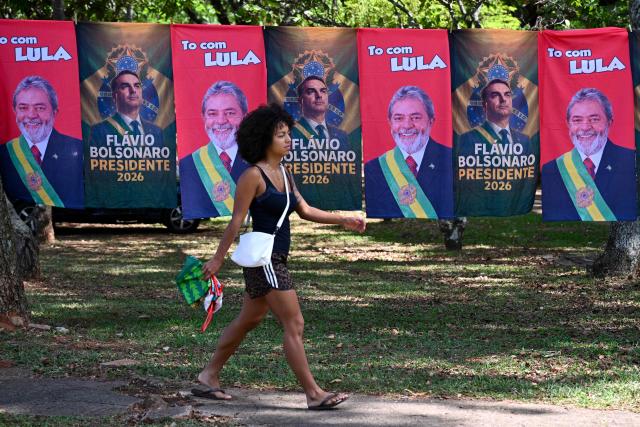 A woman walks past flags and towels bearing the images of Brazil's President and presidential candidate Luiz Inacio Lula da Silva and right-wing senator and presidential candidate Flavio Bolsonaro for sale in a sytreet in Brasiilia on October 22, 2026. Lula da Silva and Bolsonaro head to a runoff election on October 25, a race that has become mired in allegations of social media malfeasance. (Photo by Evaristo Sa / AFP)