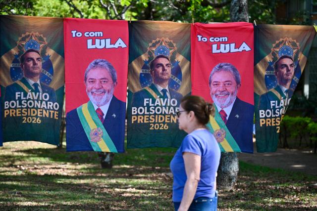 A woman walks past flags and towels bearing the images of Brazil's President and presidential candidate Luiz Inacio Lula da Silva and right-wing senator and presidential candidate Flavio Bolsonaro for sale in a street in Brasiilia on October 22, 2026. Lula da Silva and Bolsonaro head to a runoff election on October 25, a race that has become mired in allegations of social media malfeasance. (Photo by Evaristo Sa / AFP)
