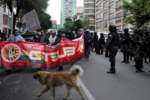 Teachers hold a banner of the Confederation of Urban Education Workers of Bolivia (CTEUB) as they march during a protest against the government's economic and educational policies in La Paz on October 22, 2026, while police officers stand nearby. (Photo by Aizar RALDES / AFP)