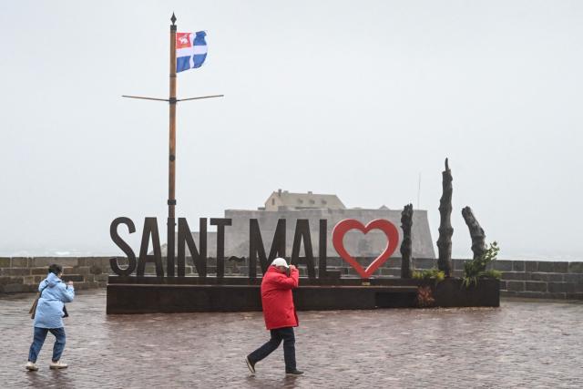 Pedestrians walks past a set of letters reading "Saint Malo" along the sea during the Austen storm in Saint Malo, western France on October 23, 2026 before the start of the 13th Route du Rhum Destination Guadeloupe. From Saint-Malo in Brittany to Pointe-a-Pitre in Guadeloupe, over 100 skippers across 6 classes will take the start on November 1, 2026, in this solo transatlantic sailing race. (Photo by Sebastien Salom-Gomis / AFP)