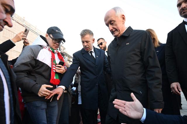 Hungarian President Andras Baka (R) is helped to step downstairs by Hungarian Prime Minister Peter Magyar (C) as they meet with visitors in front of the parliament building in Budapest on October 23, 2026 during a flag-hosting ceremony to mark the 70th anniversary of the Hungarian Revolution of 1956, also known as the Hungarian Uprising against the Soviet communist regime. (Photo by Attila KISBENEDEK / AFP)