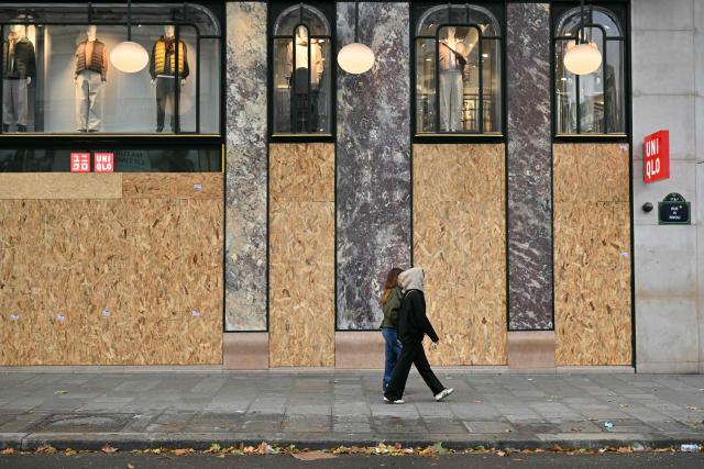 Pedestrians walk past plywood boards on a Uniqlo shop's glass windows ahead of a demonstration in favor of purchasing power, the protection of public services, and fiscal and social justice, in Paris on October 24, 2026 as part of a nationwide call for mobilisation bringing together former members of the Yellow Vest movement, labor unions, high school and university students. Protests by high school students over learning conditions sparked a movement that has since spread nationwide, fuelled by broader anger over the rising cost of living. About 70,000 security personnel will be deployed on October 24, 2026, Interior Minister said at a news conference, warning that calls for violence and looting were circulating on social media. (Photo by NICOLAS TUCAT / AFP)