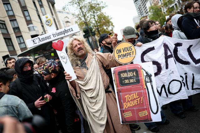 A protester holds a cross reading "Love Will Triumph" and a placard depicting a gasoline pump, reading "They're draining you dry" during a demonstration in favor of purchasing power, the protection of public services, and fiscal and social justice, in Paris on October 24, 2026 as part of a nationwide call for mobilisation bringing together former members of the Yellow Vest movement, labor unions, high school and university students. Protests by high school students over learning conditions sparked a movement that has since spread nationwide, fuelled by broader anger over the rising cost of living. About 70,000 security personnel will be deployed on October 24, 2026, Interior Minister said at a news conference, warning that calls for violence and looting were circulating on social media. (Photo by Sameer AL-DOUMY / AFP)