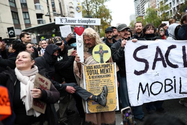 A protester (C) holds a cross reading "Love Will Triumph" as a bystander is showing their electronic ankle monitor during a demonstration in favor of purchasing power, the protection of public services, and fiscal and social justice, in Paris on October 24, 2026 as part of a nationwide call for mobilisation bringing together former members of the Yellow Vest movement, labor unions, high school and university students. Protests by high school students over learning conditions sparked a movement that has since spread nationwide, fuelled by broader anger over the rising cost of living. About 70,000 security personnel will be deployed on October 24, 2026, Interior Minister said at a news conference, warning that calls for violence and looting were circulating on social media. (Photo by Sameer AL-DOUMY / AFP)