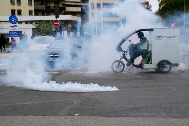 This photograph shows a car and a cyclist amid a cloud of tear gas and next to a fuming tear gas pellet during a demonstration in favor of purchasing power, the protection of public services, and fiscal and social justice, in Lyon, central France on October 24, 2026 as part of a nationwide call for mobilisation bringing together former members of the Yellow Vest movement, labor unions, high school and university students. Protests by high school students over learning conditions sparked a movement that has since spread nationwide, fuelled by broader anger over the rising cost of living. About 70,000 security personnel will be deployed on October 24, 2026, Interior Minister said at a news conference, warning that calls for violence and looting were circulating on social media. (Photo by Olivier CHASSIGNOLE / AFP)