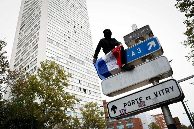 A protester holding a French national flag sits atop a public road sign during a demonstration in favor of purchasing power, the protection of public services, and fiscal and social justice, in Paris on October 24, 2026 as part of a nationwide call for mobilisation bringing together former members of the Yellow Vest movement, labor unions, high school and university students. Protests by high school students over learning conditions sparked a movement that has since spread nationwide, fuelled by broader anger over the rising cost of living. About 70,000 security personnel will be deployed on October 24, 2026, Interior Minister said at a news conference, warning that calls for violence and looting were circulating on social media. (Photo by Sameer AL-DOUMY / AFP)