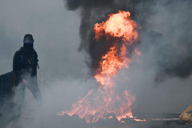 A protester stands next to a fire near Place de la Bourse, in a protest ban zone, as part of a demonstration in favor of purchasing power, the protection of public services, and fiscal and social justice, in Bordeaux, western France on October 24, 2026 as part of a nationwide call for mobilisation bringing together former members of the Yellow Vest movement, labor unions, high school and university students. Protests by high school students over learning conditions sparked a movement that has since spread nationwide, fuelled by broader anger over the rising cost of living. About 70,000 security personnel will be deployed on October 24, 2026, Interior Minister said at a news conference, warning that calls for violence and looting were circulating on social media. (Photo by Philippe LOPEZ / AFP)