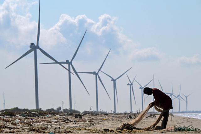 This photograph taken on October 3, 2026 shows a fisherman working on fishing nets at a beach near a wind power plant in Sri Lanka's Mannar Peninsula. The government aims to generate 70 percent of the country's electricity from renewable sources by 2030, including wind power. But it has frozen new projects in Mannar to ease local opposition. Since they appeared six years ago, the Mannar wind turbines have been blamed for a host of problems -- from dwindling catches to floods, fuelling opposition to green energy projects as fuel costs soar. (Photo by Ishara S. KODIKARA / AFP)