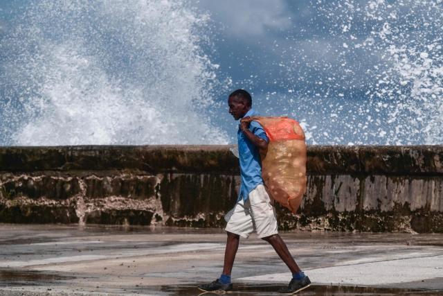 A person carries a sack of recyclable materials along the Malecon in Havana, Cuba, on October 26, 2026, as waves crash against the seawall in the background. (Photo by YAMIL LAGE / AFP)