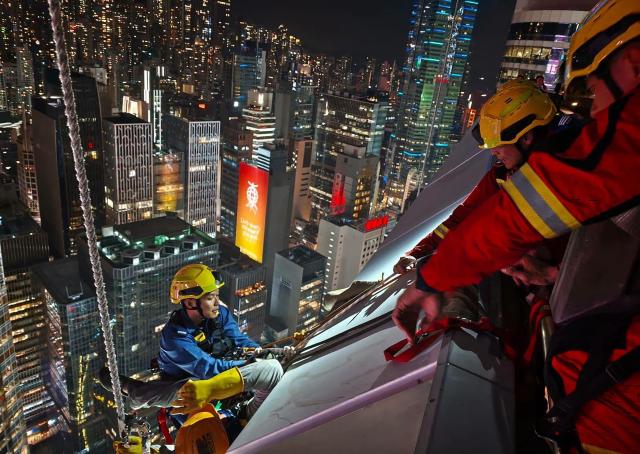 This handout photo taken on October 26, 2026 and released on October 27 by the Hong Kong Fire Services Department shows firefighters rescuing maintenance workers who were dangling on safety harnesses after their working platform malfunctioned on the outside of Jardine House in the Central business district in Hong Kong. (Photo by Handout / HONG KONG FIRE SERVICES DEPARTMENT / AFP)