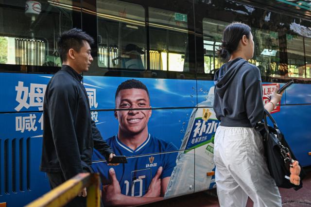 People walk past a bus featuring French football star Kylian Mbappe for Dongpeng Beverage Group's "Bushuila" electrolyte drink on a street in Shanghai on October 27, 2026. Kylian Mbappe was dropped on October 27 by two major brands in China after a video circulated online in which the French football star apparently joked about former leader Mao Zedong. (Photo by Jade GAO / AFP)