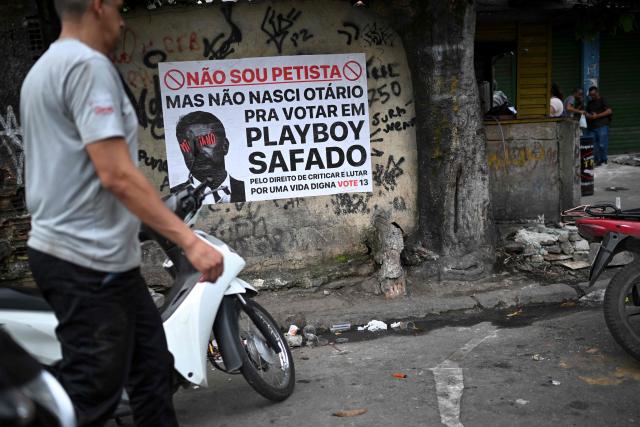 This view shows a banner depicting Brazilian Senator Flavio Bolsonaro with text in Portuguese that reads, “I’m not a Workers’ Party supporter, but I’m not dumb enough to vote for a douchebag playboy. For the right to criticize and fight for a better life, vote 13,” on a wall at the entrance to the Vila Cruzeiro favela, part of the Penha Complex, in Rio de Janeiro, Brazil, on October 28, 2026. (Photo by MAURO PIMENTEL / AFP)