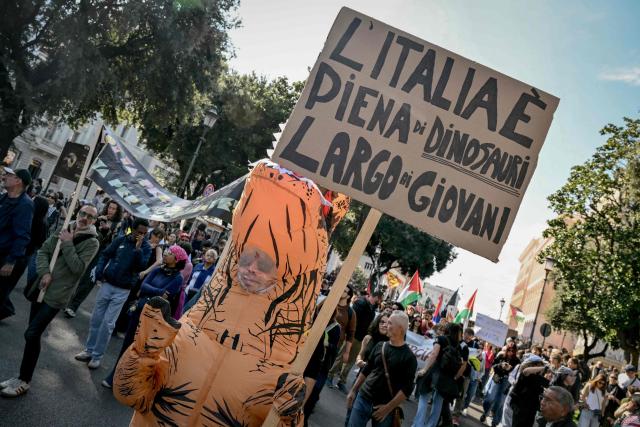 A person wearing a dinosaur costume holds a placard reading in Italian "Italy is full of young people who are as clueless as dinosaurs" during a demonstration part of a nationwide day of strikes and protests as school and university students demand more funding for schools and support for housing in Rome on October 30, 2026. Students have been protesting for weeks, setting up tent camps and occupying schools to make their demands heard, matching similar movements across the border in France. The Italian protests have taken place alongside those of truck drivers and farmers over the spike in fuel prices and the cost of living, as well as rising defence spending. (Photo by Alberto PIZZOLI / AFP)
