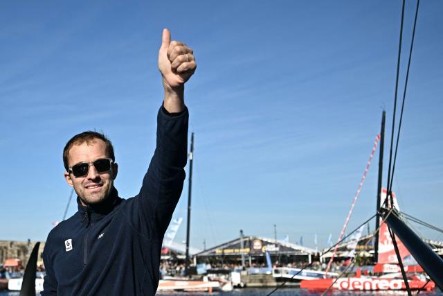 British sailor Sam Goodchild waves to the public aboard his "Macif Sante Prevoyance" IMOCA Class as he prepares to pass through the Naye lock of Saint-Malo's harbour, to moor off the coast of Saint-Malo northwestern France on October 31, 2026 on the eve of the start of the Route du Rhum solo sailing race from Saint-Malo, to Pointe-a-Pitre in the French overseas island of Guadeloupe. (Photo by Loic VENANCE / AFP)