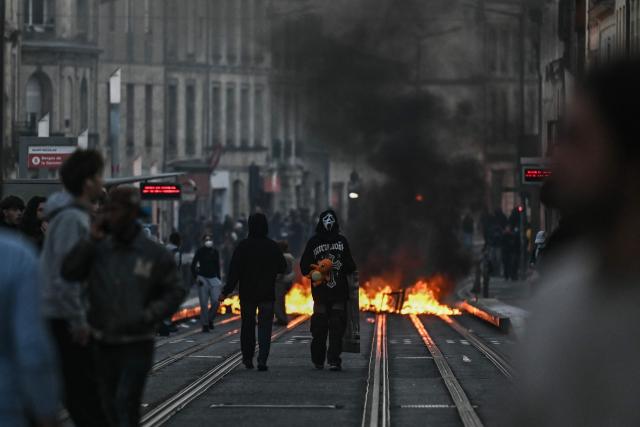 A protester with a mask referencing Ghostface, a character from the Scream movie franchise stands next to a street fire on tram rails and holds a doll referencing Pokemon character Charmander during nationwide demonstrations over the rising cost of living and inequality organised by Yellow Vest movement, labour unions and high school and university students, in Bordeaux, southwestern France on October 31, 2026. "Exceptional" security deployment with 70,000 police officers and gendarmes mobilized and an alarmist tone: for Act III of the protests against the high cost of living, right in the middle of Halloween festivities, the authorities fear a repeat of the October 17 nightmare. France has been rocked by widespread anger over the rising cost of living and inequality. (Photo by Christophe ARCHAMBAULT / AFP)