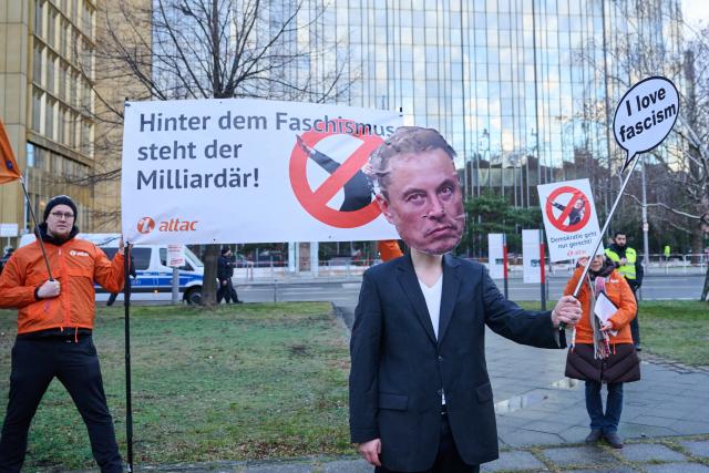 27 January 2025, Berlin: An Attac activist is seen wearing mask depicting businessman and political figure Elon Musk, while others hold a banner reading "Behind fascism is the billionaire!", during a demonstration in front of the Axel Springer skyscraper in conjunction with an economic summit by the German newspaper "Die Welt". Photo: Annette Riedl/dpa