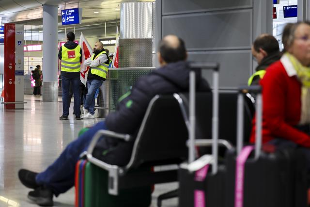 10 March 2025, North Rhine-Westphalia, Duesseldorf: Airport employees wearing high-visibility vests from the Verdi union stand in the departures area at Duesseldorf Airport as travellers wait for their flights. The Verdi union has called for a 24-hour warning strike by public service and ground handling workers at eleven airports on Monday. Photo: Christoph Reichwein/dpa