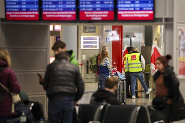 10 March 2025, North Rhine-Westphalia, Duesseldorf: Airport employees wearing high-visibility vests from the Verdi union stand in the departures area at Duesseldorf Airport as travellers wait under the display boards. The Verdi union has called for a 24-hour warning strike by public service and ground handling workers at eleven airports on Monday. Photo: Christoph Reichwein/dpa