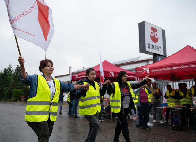 22 May 2025, Baden-Württemberg, Murr: Three strikers are dancing, one holding a flag of the NGG trade union. A warning strike is taking place at the Birtat kebab factory in Murr. The strike was called by the Food, Beverages and Catering Union (NGG) because previous negotiations on the first collective bargaining agreement have been unsuccessful. Photo: Markus Lenhardt/dpa