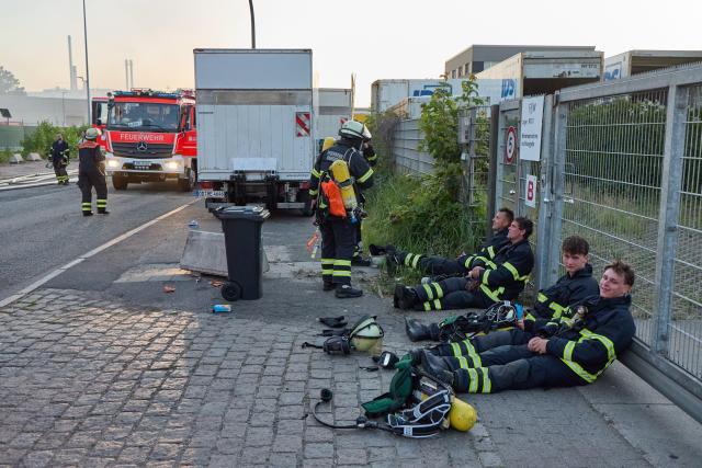01 July 2025, Hamburg: Exhausted firefighters rest by the roadside as they battle a fire at a waste recycling company's warehouse in Hamburg-Billbrook. No injuries have been reported and no one is currently in danger, according to the fire department's situation center. It remains unclear what was being stored in the warehouse. Photo: Georg Wendt/dpa