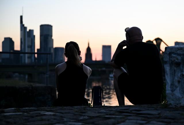 01 July 2025, Hesse, Frankfurt/Main: A woman and a man sit on the banks of the River Main, enjoying the view of the Frankfurt skyline at sunset on a hot tropical evening. Photo: Arne Dedert/dpa