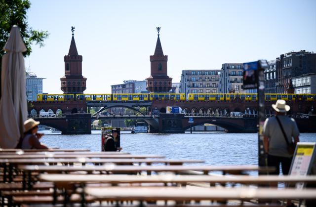 30 June 2025, Berlin: Visitors to Berlin sit and stand along the Spree near the East Side Gallery, with the Oberbaum Bridge in the background. Photo: Bernd von Jutrczenka/dpa