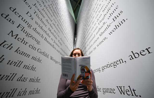 15 October 2025, Hesse, Frankfurt_Main: Ann-Kristin Mueller holds the novel "Die Passantin" by Nina George between the pages of the largest book at the Frankfurt Book Fair at the Kein & Aber publishing house stand. Photo: Arne Dedert/dpa