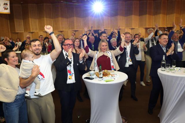 26 October 2025, Bavaria, Munich: German canoeist Sideris Tasiadis (2nd L) and former skier Hilde Gerg (C) celebrate with other visitors after the first projections on the result of the referendum on the Olympic bid were announced at the Haus des Sports. According to Munich's Lord Mayor Reiter, well over 60 percent voted in favor of the Bavarian capital's bid to host the Summer Games and Paralympic Games in 2036, 2040 or 2044. Photo: Karl-Josef Hildenbrand/dpa