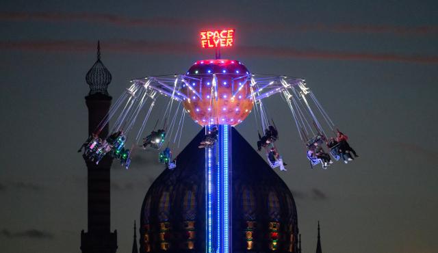 30 October 2025, Saxony, Dresden: The 'Space Flyer' ride, a type of chain carousel in which not only the gondolas but also the entire tower rotate, is illuminated with colorful lights in the evening at the autumn festival on the Pieschner Allee fairground. The Yenidze, a former cigarette factory building, can be seen in the background. Photo: Robert Michael/dpa